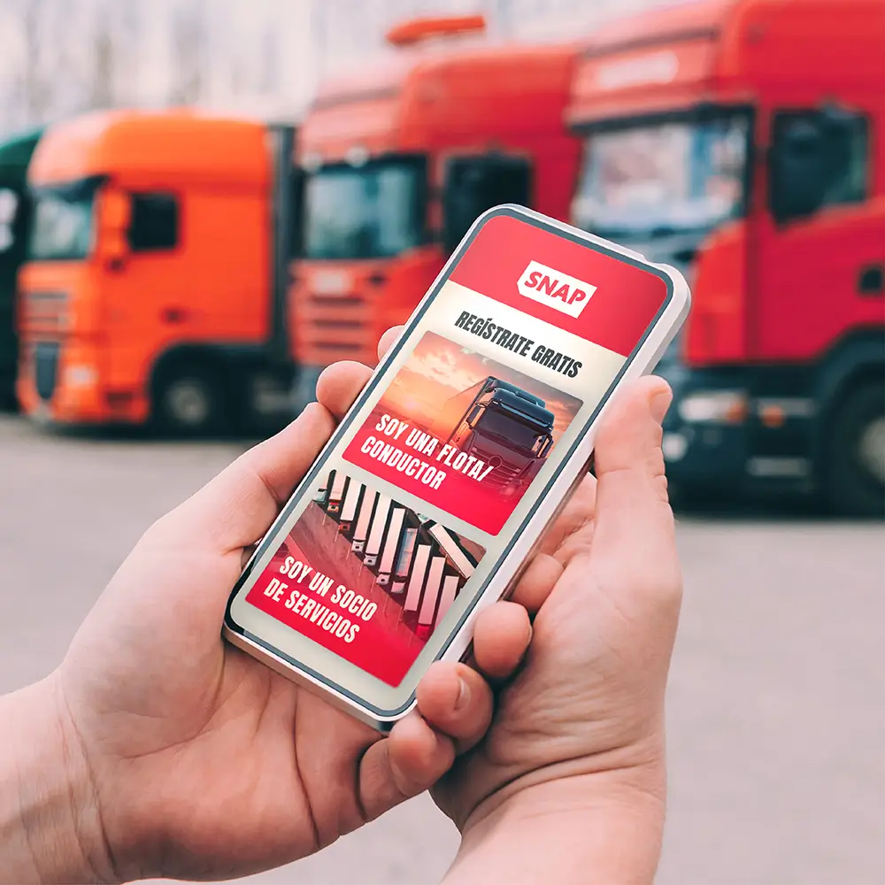 Mano sosteniendo un móvil con la app Vrio-SNAP mostrando opciones para registrarse gratis y gestionar parkings seguros para camiones y socios de servicio.